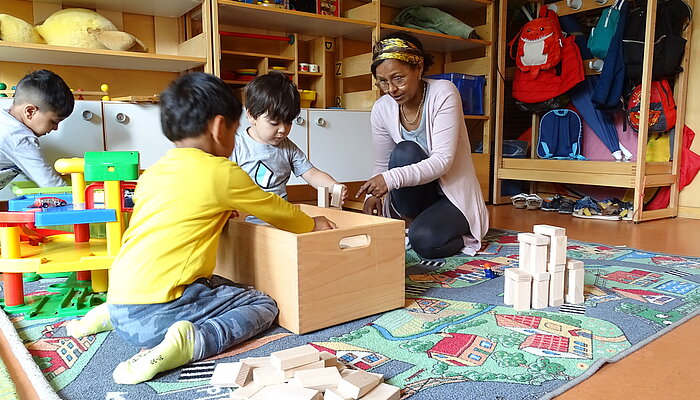 Eine Frau holt mit zwei Jungen Holzbausteine aus einer Kiste, im Hintergrund spielt ein weiterer Junge