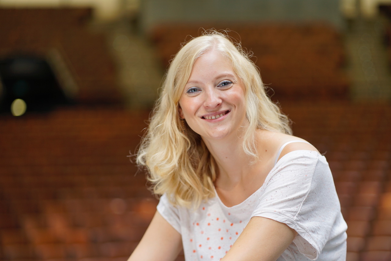 Eine blonde Frau sitzt in einem Saal und lächelt in die Kamera. Es ist Fenja Grieshammer, die sich ehrenamtlich im Mehrgenerationenhaus Wunsiedel engagiert.