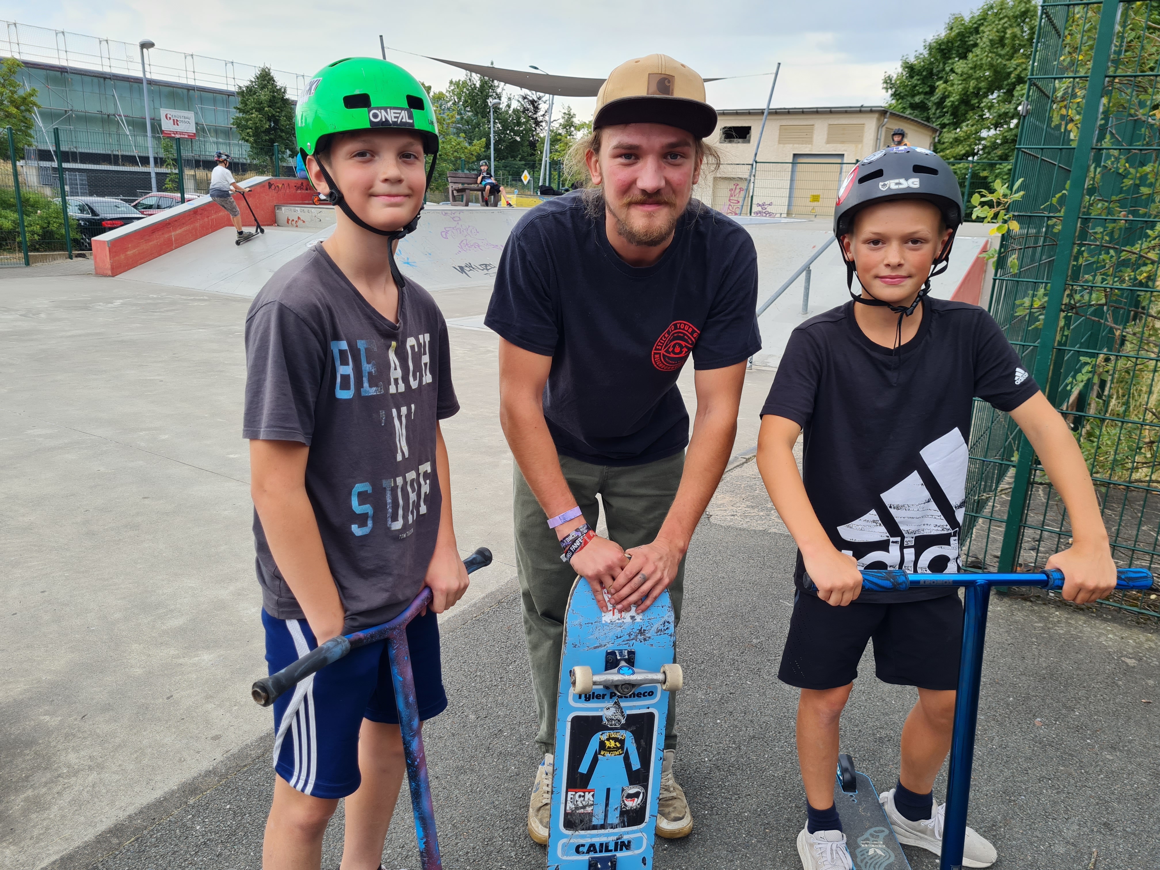 Drei Jungs tragen Helme, stehen auf ihren Rollern und lächeln.