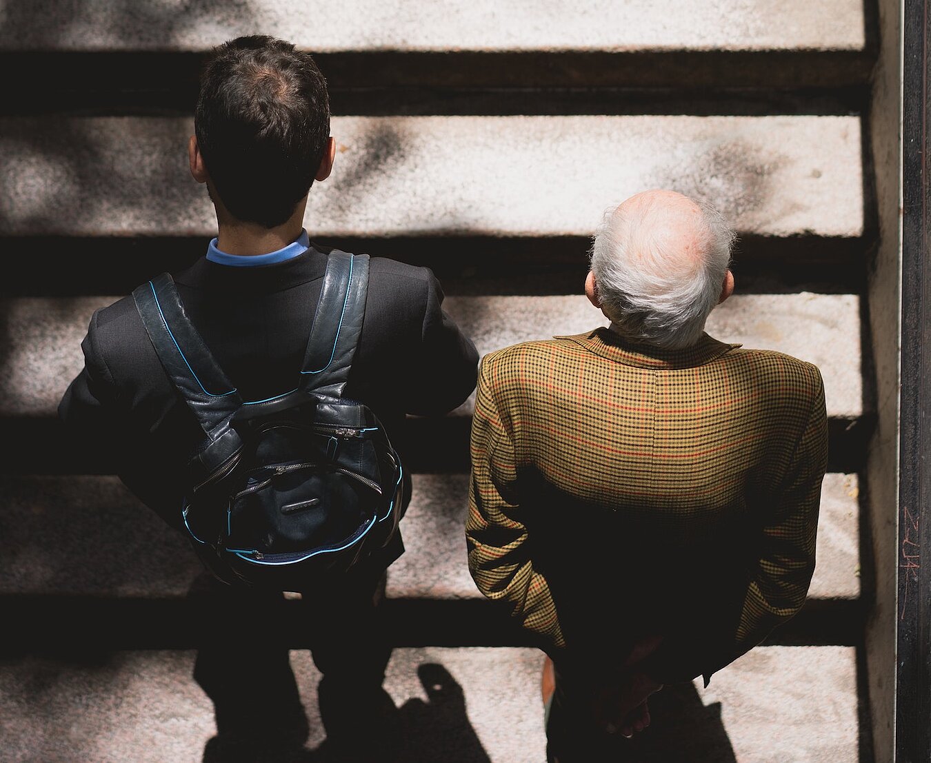 Ein junger Mann mit Rucksack und ein älterer Mann mit grauen Haaren und Glatze gehen eine Treppe hoch, Perspektive von oben auf die Köpfe