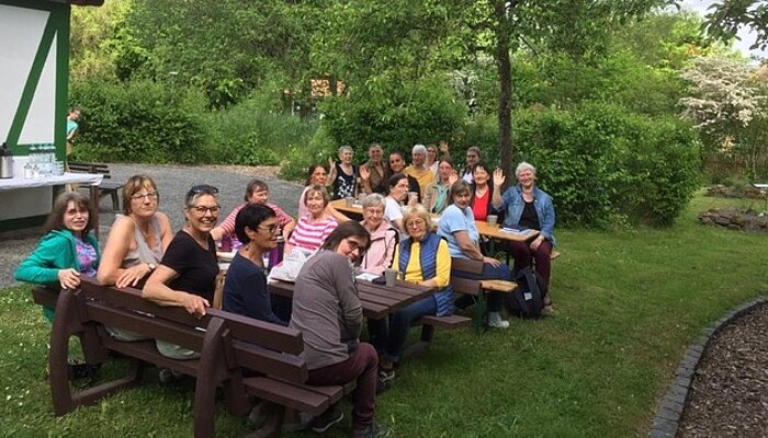 Viele Frauen sitzen auf Holzbänken an Holztischen in einem Garten.