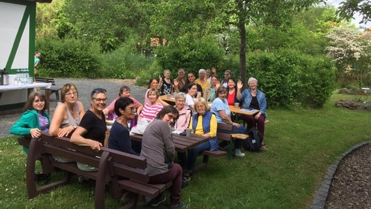 Viele Frauen sitzen auf Holzbänken an Holztischen in einem Garten.