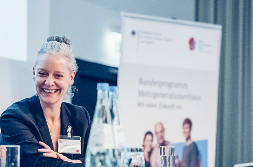 Foto von Maren Duesberg, die auf einem Podium sitzt und lächelt, im Hintergrund ein Roll-Up des Bundesprogramms Mehrgenerationenhaus.