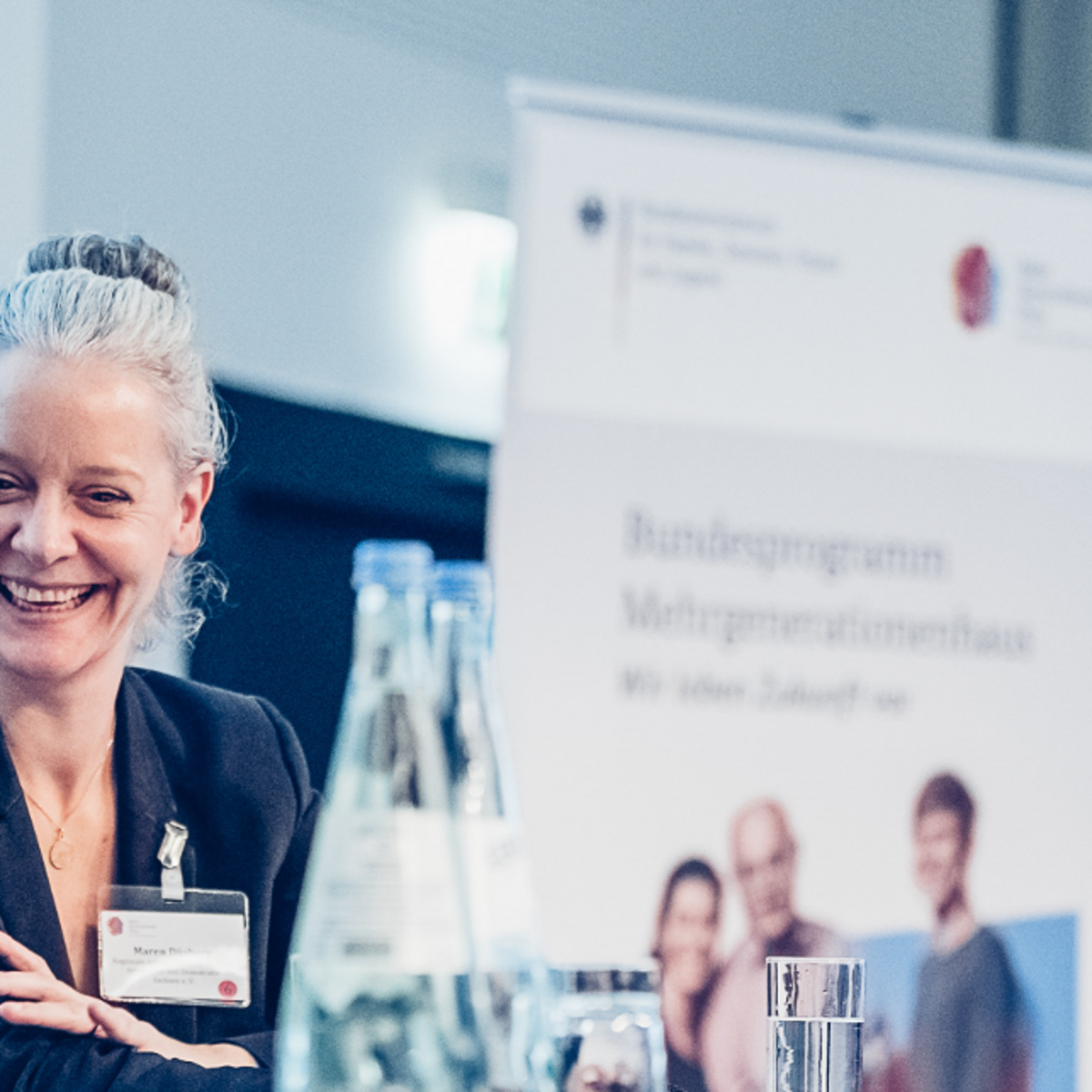 Foto von Maren Duesberg, die auf einem Podium sitzt und lächelt, im Hintergrund ein Roll-Up des Bundesprogramms Mehrgenerationenhaus.