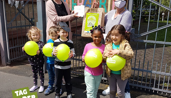 Fünf Kinder mit Luftballons stehen vor dem Kindergarten. Im Hintergrund zwei Frauen mit einer Tüte mit den 90 Postkarten