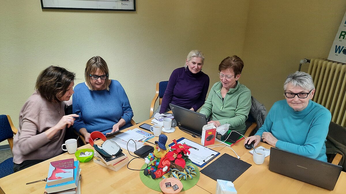 Fünf Frauen sitzen an einem Tisch und schauen auf Handybildschirme und Laptops
