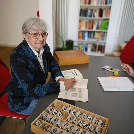 Frau mit grauen Haaren, Brille und blauem Blazer sitzt an einem Tisch, auf dem ein Kasten mit Buchstabenplatten steht