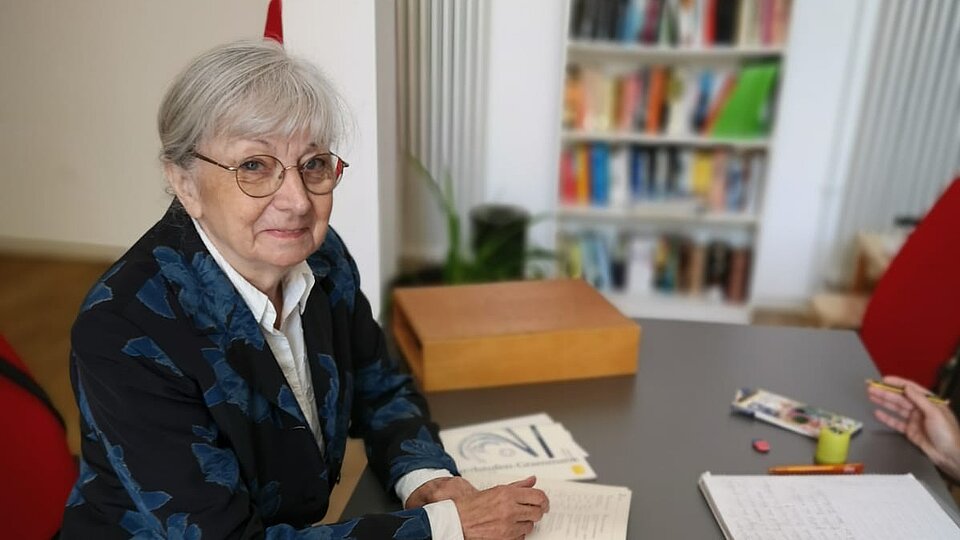 Frau mit grauen Haaren, Brille und blauem Blazer sitzt an einem Tisch, auf dem ein Kasten mit Buchstabenplatten steht