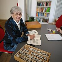 Frau mit grauen Haaren, Brille und blauem Blazer sitzt an einem Tisch, auf dem ein Kasten mit Buchstabenplatten steht
