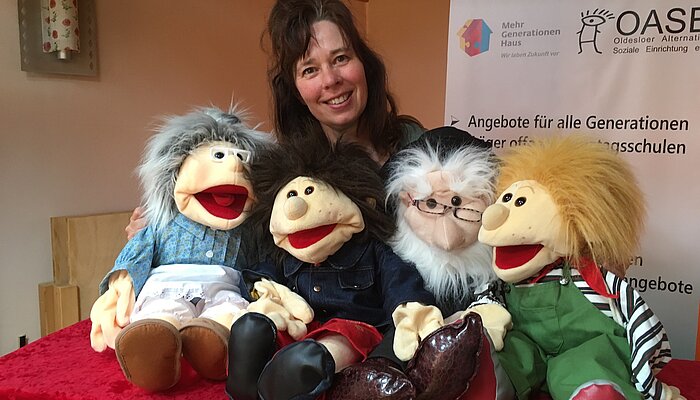 Frau steht hinter einem Tisch, auf dem vier Handpuppen sitzen