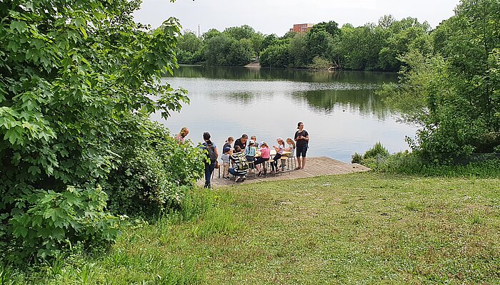 Sieben Kinder und drei Erwachsene sitzen an einem Tisch vor einem See.
