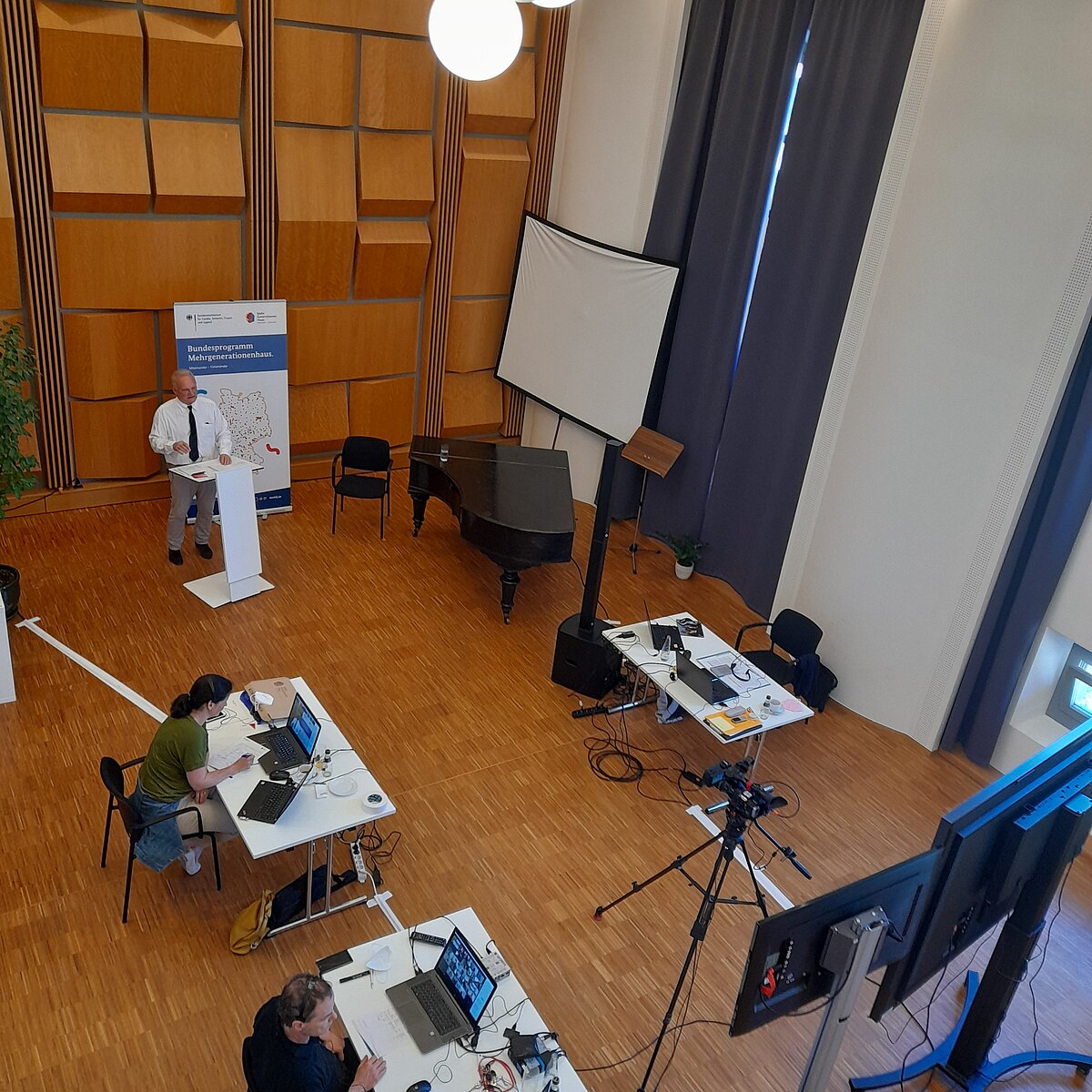 © BAFzA Blick von oben auf Aufbau des Fachtags mit Moderator am Pult und zwei Personen an Schreibtischen mit Laptops