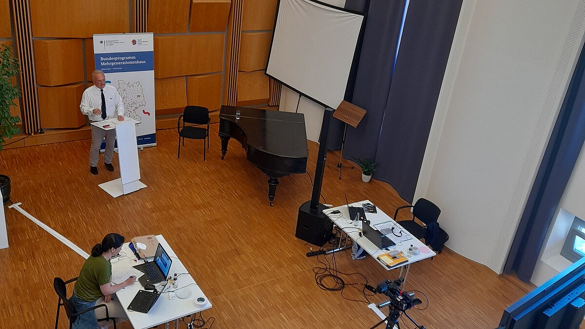 Blick von oben auf Aufbau des Fachtags mit Moderator am Pult und zwei Personen an Schreibtischen mit Laptops
