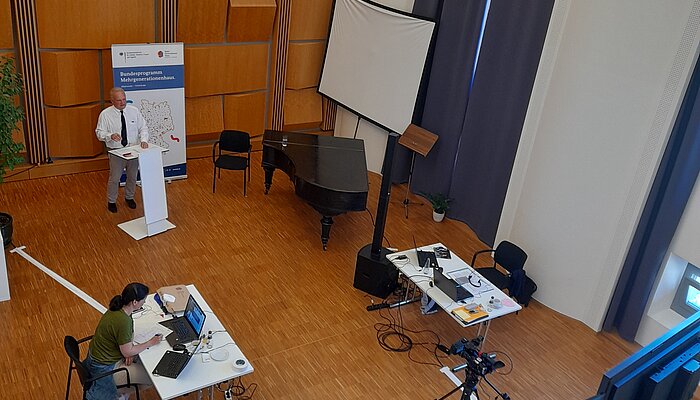 Blick von oben auf Aufbau des Fachtags mit Moderator am Pult und zwei Personen an Schreibtischen mit Laptops