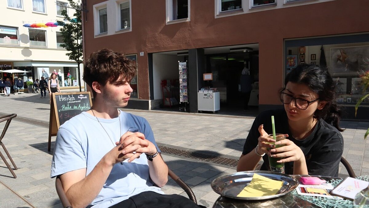 Eine junge Frau und ein junger Mann sitzen an einem Bistrotisch und trinken Smoothies.
