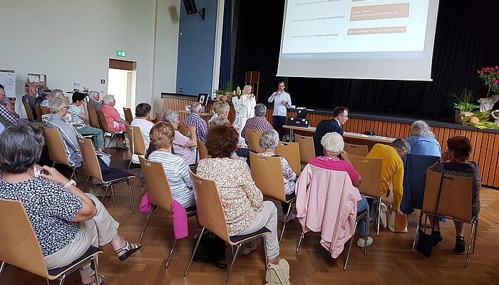 Ältere Menschen sitzen in Stuhlreihen in einer Aula und verfolgen einen Vortrag.