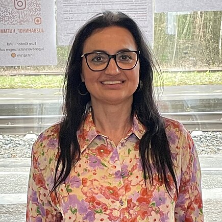 Eine Frau mit schwarzen langen Haaren und Brille lehnt an einer Fensterbank und lächelt in die Kamera.