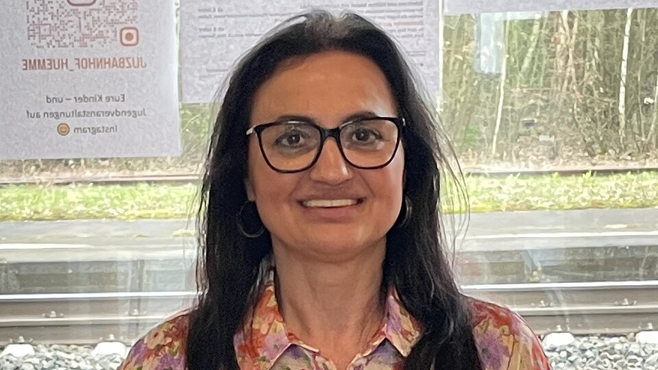 Eine Frau mit schwarzen langen Haaren und Brille lehnt an einer Fensterbank und lächelt in die Kamera.