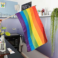 Hand hält von links eine Regenbogenfahne ins Bild, im Hintergrund steht die Bar eines Cafés.