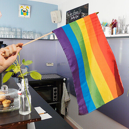 Hand hält von links eine Regenbogenfahne ins Bild, im Hintergrund steht die Bar eines Cafés.