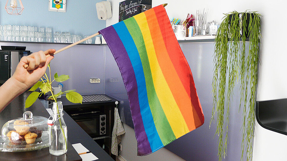 Hand hält von links eine Regenbogenfahne ins Bild, im Hintergrund steht die Bar eines Cafés.
