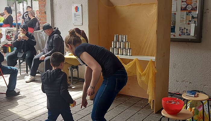 Eine Frau zeigt auf den Boden, ein Kind hält einen Ball in der Hand, auf einem Tisch stehen Blechdosen in einer Pyramide