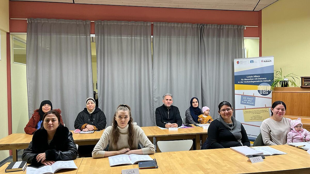 Sieben Frauen und ein Mann sitzen in zwei Reihen an Schultischen, zwei Frauen haben jeweils ein Baby auf dem Schoß