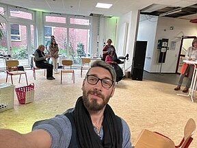 Selfie von Tobias Wiedemann vor einem großen Raum mit sechs Menschen im Hintergrund