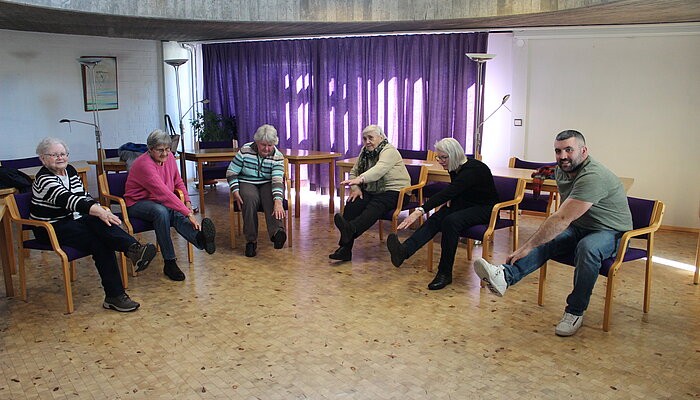 Fünf ältere Frauen und ein Mann sitzen auf Stühlen und strecken ein Bein und einen Arm aus