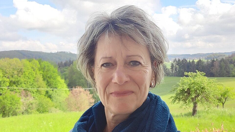 Eine Frau mit kurzen, grauen Haaren lacht in die Kamera. Hinter ihr ist ein Feld mit Bäumen zu sehen.