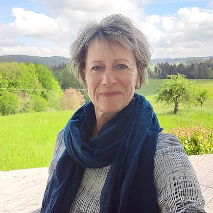 Eine Frau mit kurzen, grauen Haaren lacht in die Kamera. Hinter ihr ist ein Feld mit Bäumen zu sehen.