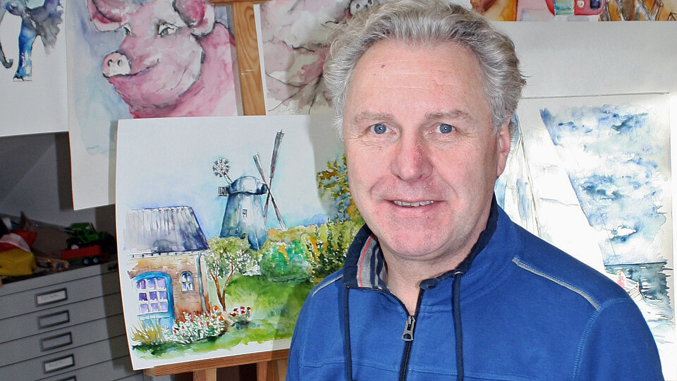 Mann sitzt mit Pinsel vor einem leeren Blatt. Im Hintergrund hängen Aquarellbilder mit unterschiedlichen Motiven.