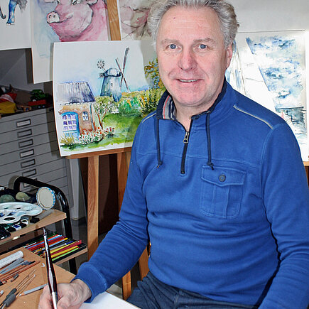 Mann sitzt mit Pinsel vor einem leeren Blatt. Im Hintergrund hängen Aquarellbilder mit unterschiedlichen Motiven.