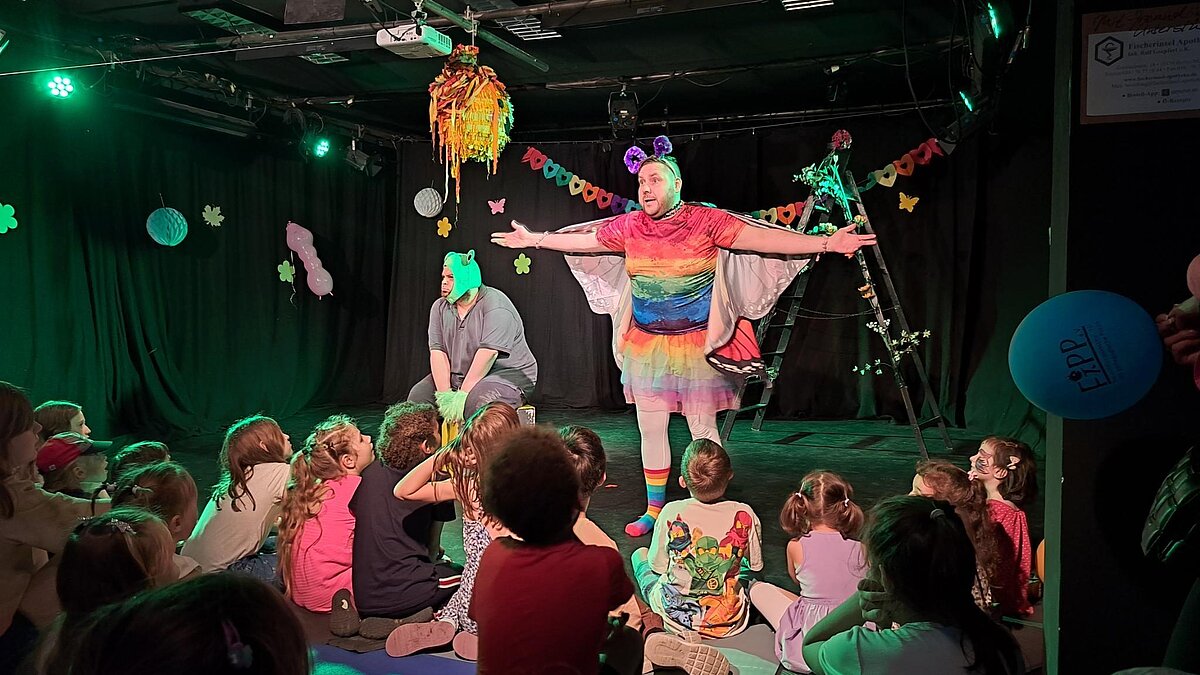 Das Bild zeigt eine Theateraufführung für Kinder. Ein Mann im Schmetterlingskostüm steht vor dem Publikum.