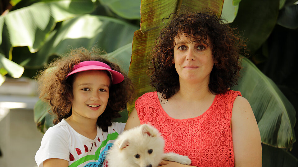 Mädchen mit braunen Locken und buntem T-Shirt neben Frau mit braunen Locken und rotem Top einen Hund auf dem Arm haltend