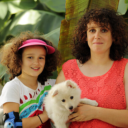 Mädchen mit braunen Locken und buntem T-Shirt neben Frau mit braunen Locken und rotem Top einen Hund auf dem Arm haltend