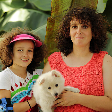 Mädchen mit braunen Locken und buntem T-Shirt neben Frau mit braunen Locken und rotem Top einen Hund auf dem Arm haltend