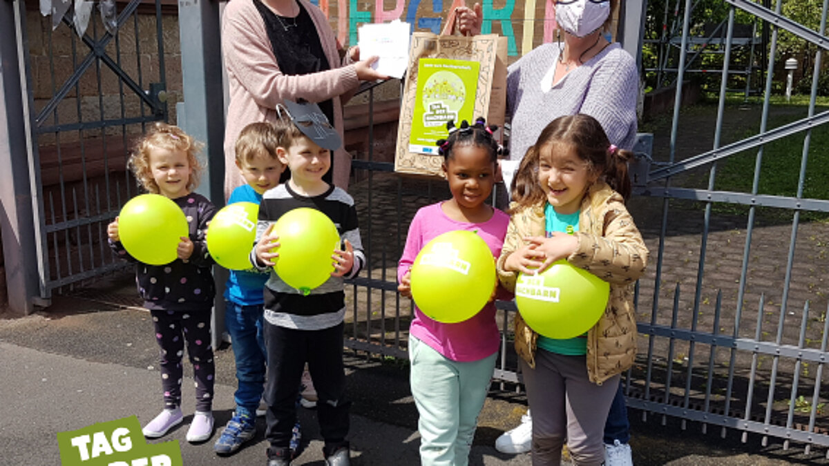 Fünf Kinder mit gelben Ballons stehen vor einem Zaun, dahinter stehen zwei Frauen, die eine Tüte hochhalten