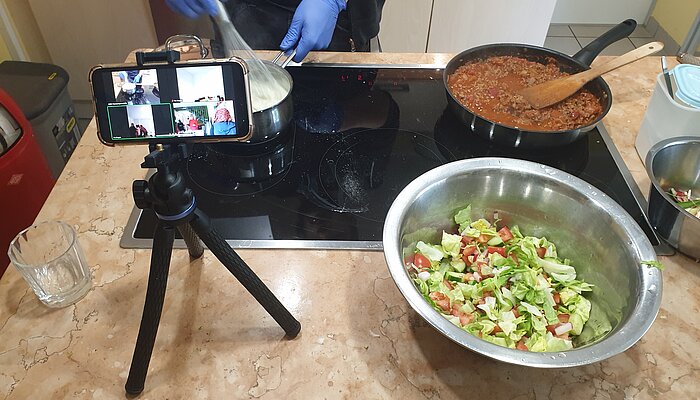 Aufnahme von oben auf einen Herd, auf dem eine Pfanne mit Hackfleisch-Soße und ein Topf mit weiter Flüssigkeit stehen, die weiße Flüssigkeit wird von zwei Händen mit Handschuhen und einem Schneebesen verrührt, vor dem Herd stehen ein Stativ mit Handy und eine Schüssel Salat