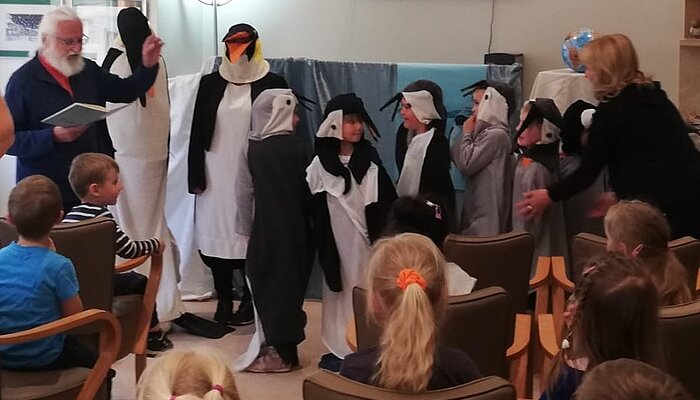 Zwei Erwachsene und sechs Kinder stehen in Pinguinkostümen vor einer Wand, ein Mann liest vor, mehrere Kinder schauen zu