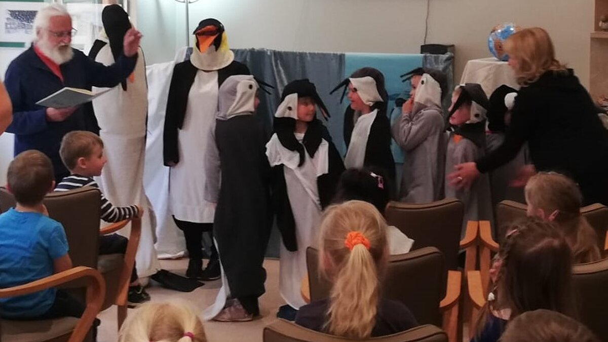 Zwei Erwachsene und sechs Kinder stehen in Pinguinkostümen vor einer Wand, ein Mann liest vor, mehrere Kinder schauen zu
