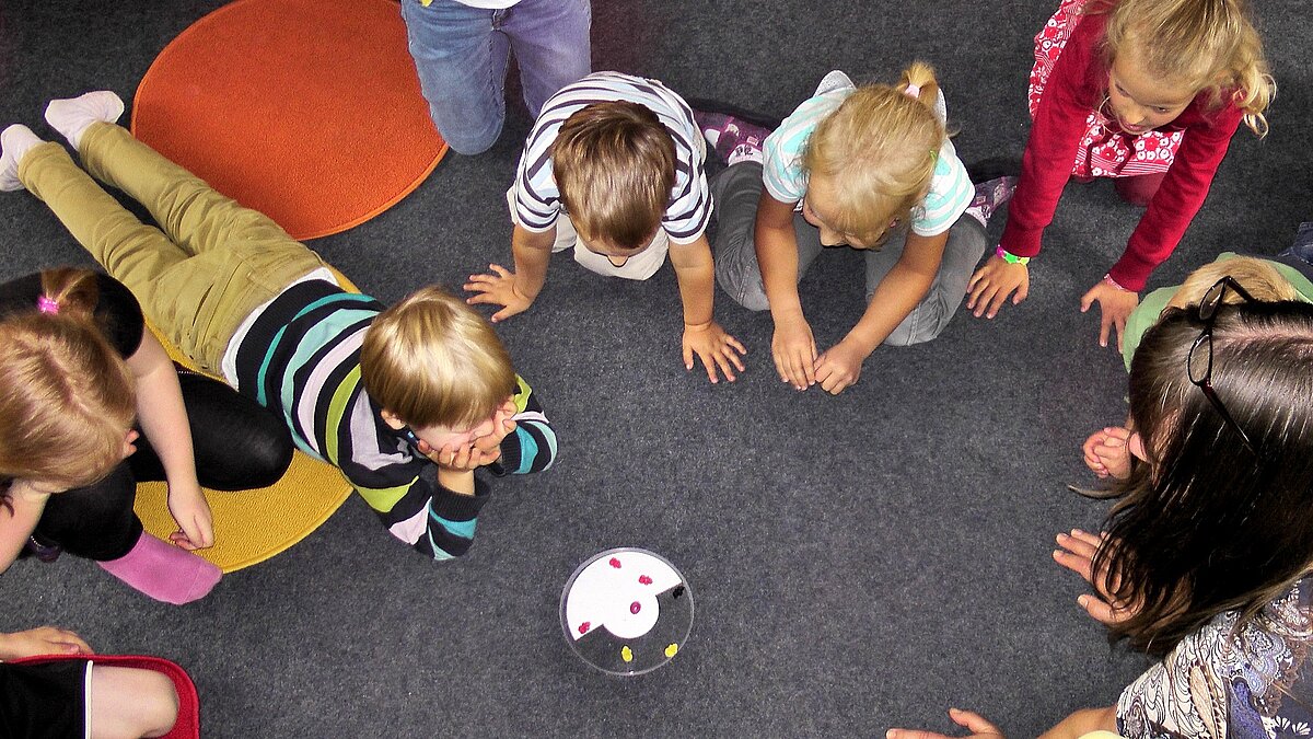 Acht Kinder und eine Frau hocken im Kreis auf dem Boden, in der Mitte steht eine Schale mit Fruchtgummi