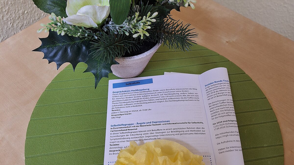 Auf einem Tisch liegen ein Platzset und eine Informationsbroschüre, dahinter steht ein Blumenstrauß