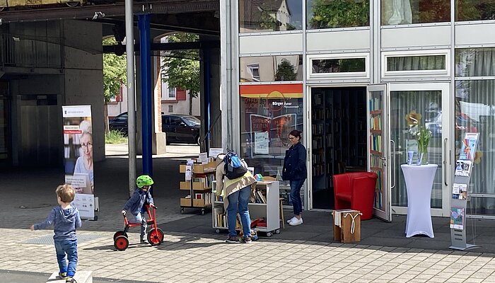 Vor einem Gebäude mit Glasfront stehen Bücherregale, zwei Frauen schauen sich die Bücher an, zwei Kinder laufen umher