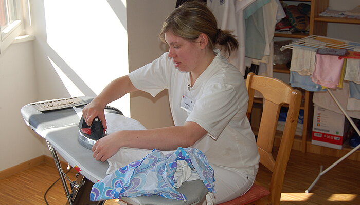 Frau in weißer Arbeitskleidung sitzt am Bügelbrett und bügelt ein weißes Hemd.