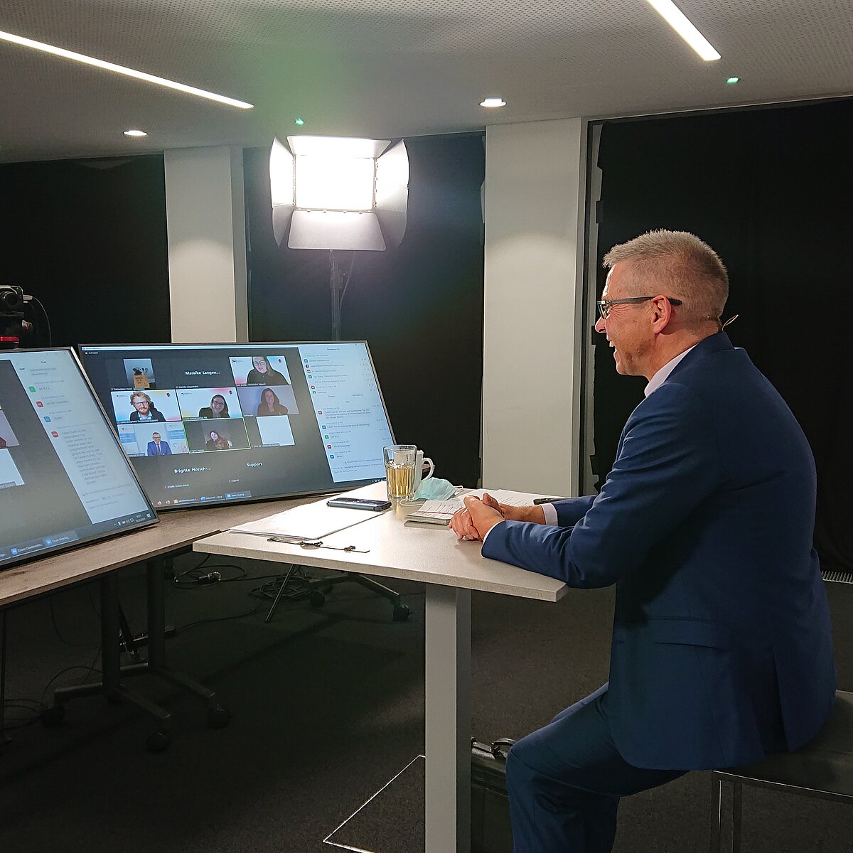 Dr. Winfried Kösters sitzt an Tisch mit Bildschirmen, auf denen Videokonferenzen  zu sehen sind