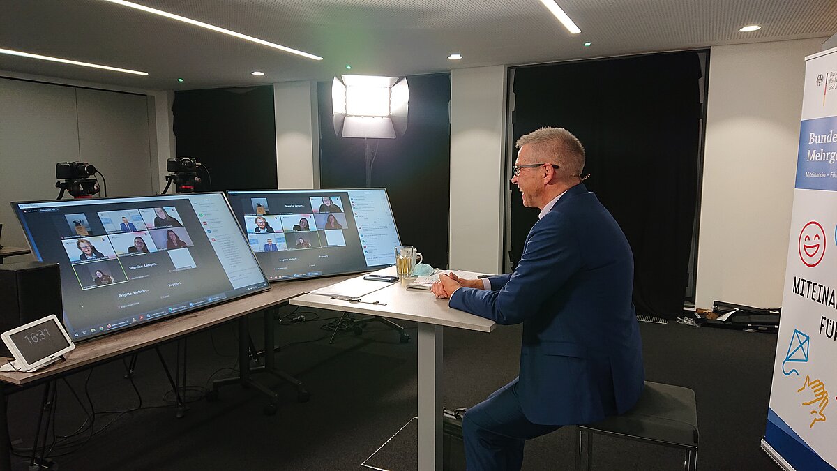 Dr. Winfried Kösters sitzt an Tisch mit Bildschirmen, auf denen Videokonferenzen  zu sehen sind