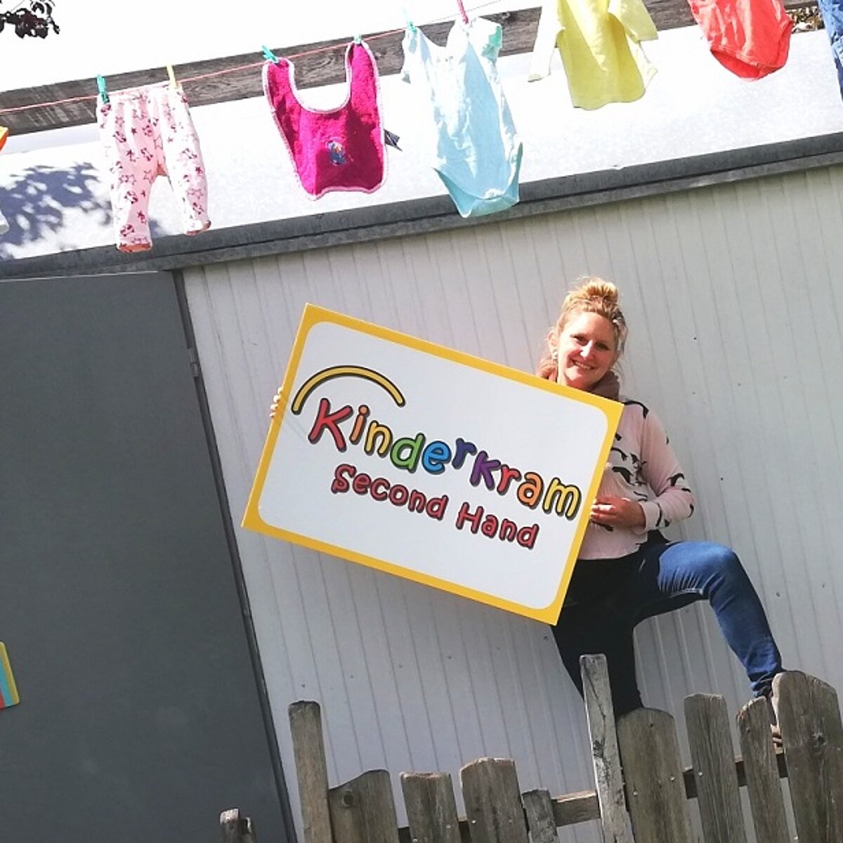 Frau steht vor einem Haus mit einem Schild in der Hand auf dem steht Kinderkrams Second Hand