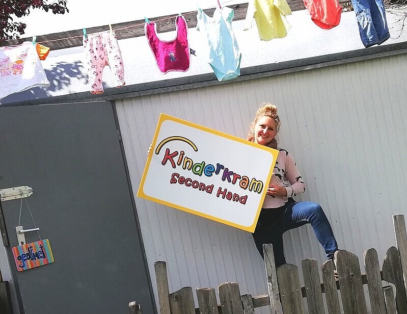 Frau steht vor einem Haus mit einem Schild in der Hand auf dem steht Kinderkrams Second Hand