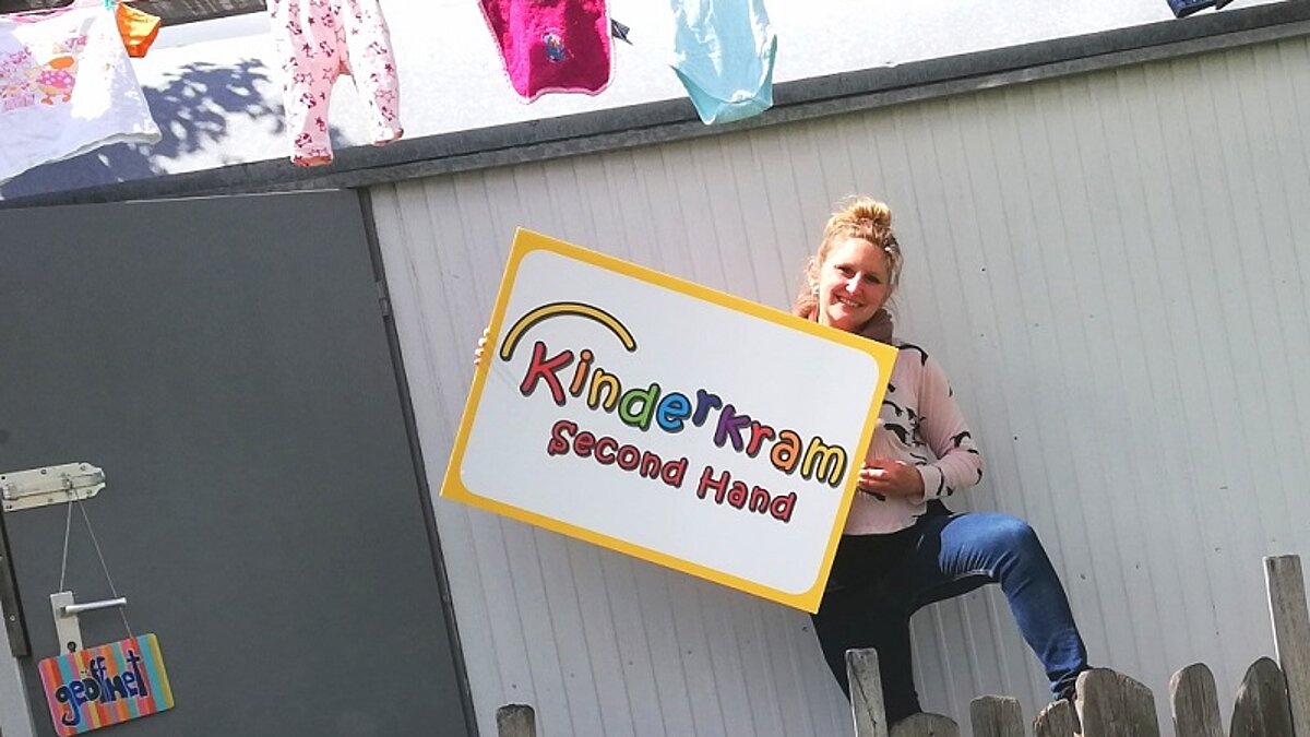 Frau steht vor einem Haus mit einem Schild in der Hand auf dem steht Kinderkrams Second Hand